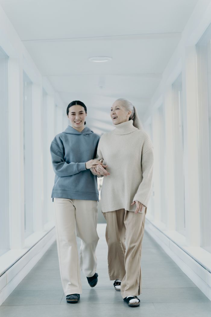 A senior woman and younger woman walking together in a bright hallway, holding hands and smiling.